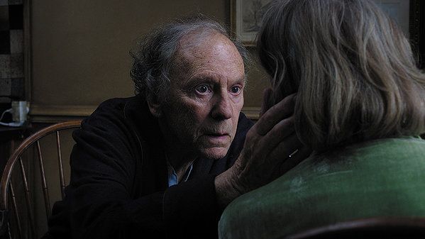 Elderly man gazing at woman, hand gently touching her face. Interior setting, soft light.
