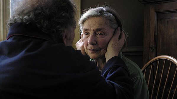 Elderly woman with concerned expression, face held gently by another, in a dimly lit room.