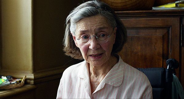 Elderly woman with gray hair and glasses speaks, in a room with a window.