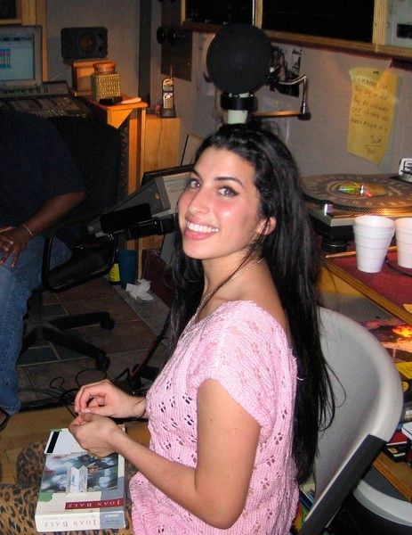 Amy Winehouse smiling in a recording studio, wearing a pink top, with a microphone overhead.