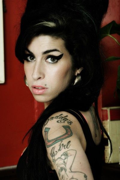 Amy Winehouse with winged eyeliner, looking at the camera, her arm displaying tattoos.