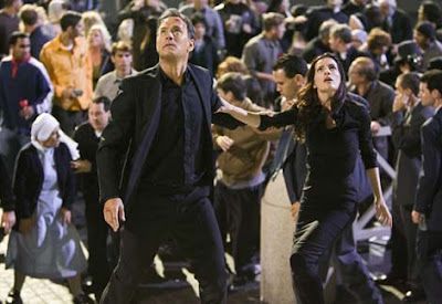 Man and woman in black, looking up, amidst a crowd of people. Outdoors, evening.