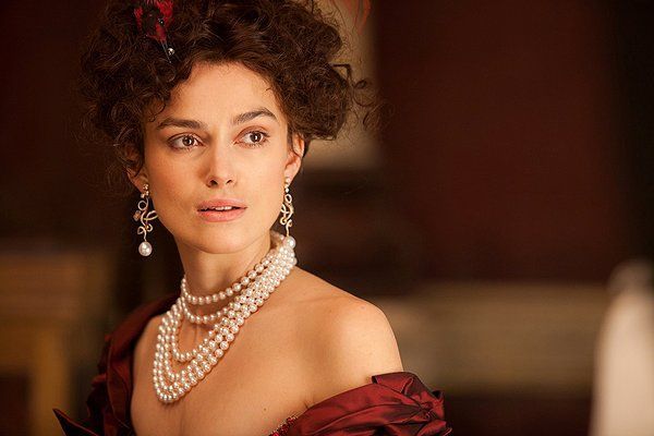 Woman in period attire with pearls, gazing off-screen, dark red dress, ornate earrings.