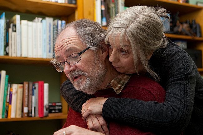 Older woman hugging older man from behind; bookshelves in background.