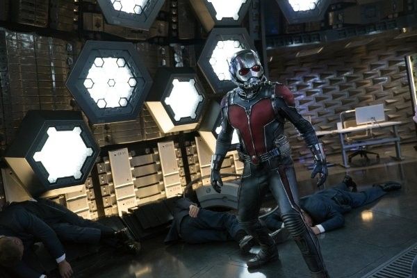 Ant-Man in his suit stands over fallen men in a metallic room, illuminated by geometric lights.