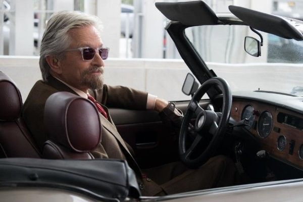 Michael Douglas in a convertible car, wearing sunglasses and a suit, looking to the side.