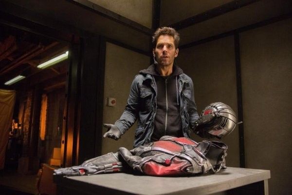 Paul Rudd holding Ant-Man helmet, standing near disassembled suit on a table in a dimly lit room.