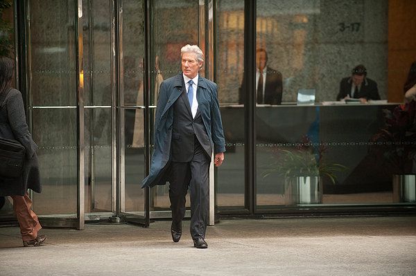 Richard Gere in a suit and coat exits a glass door of a building, walking forward.
