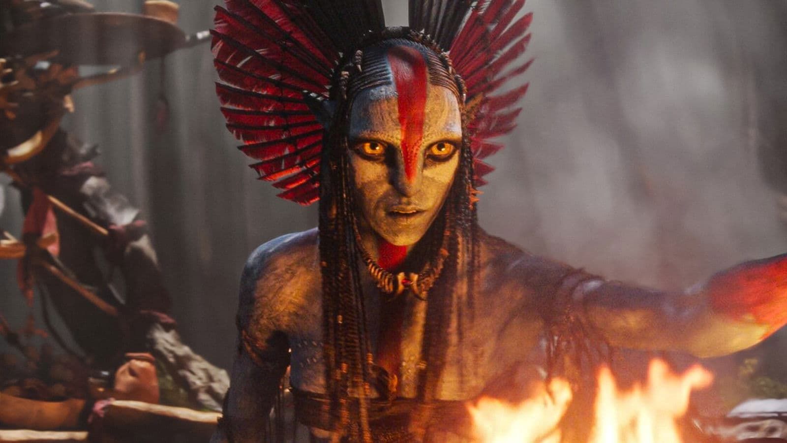 Masked figure with red feathered headdress, lit by fire in a smoky battle scene