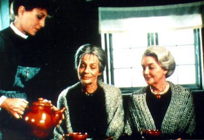 Woman pouring tea for two seated older women. Dark room, simple decor.