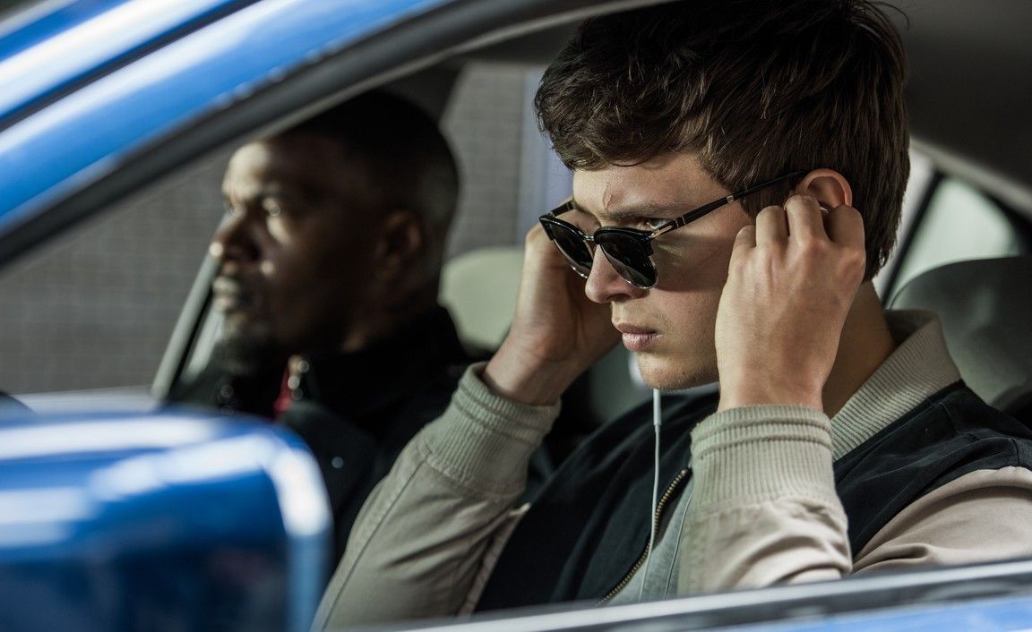 A young man in sunglasses adjusts earbuds in a car, another man looks on.