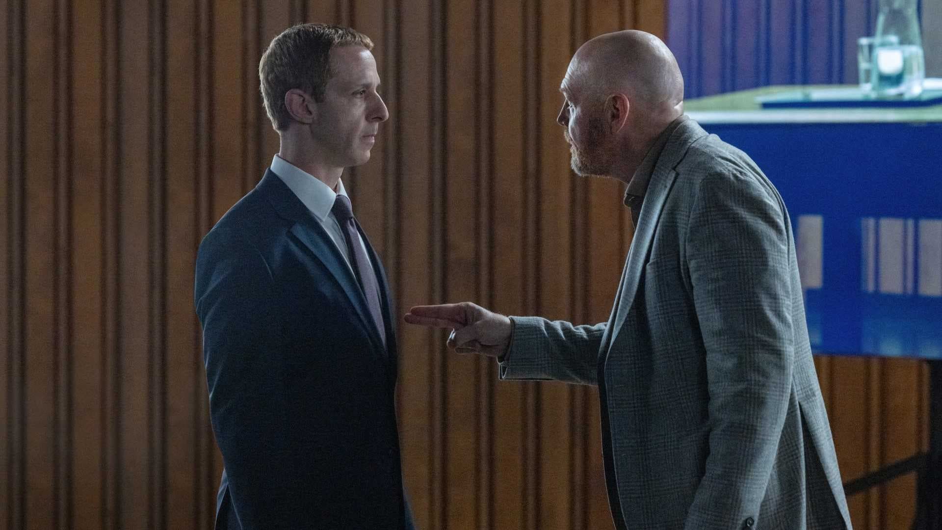 Two men in suits face off in a tense office, one pointing a finger at the other.