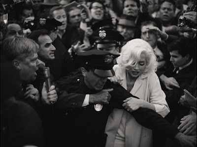 Marilyn Monroe, surrounded by a crowd and police, is being led through the throng. Black and white.