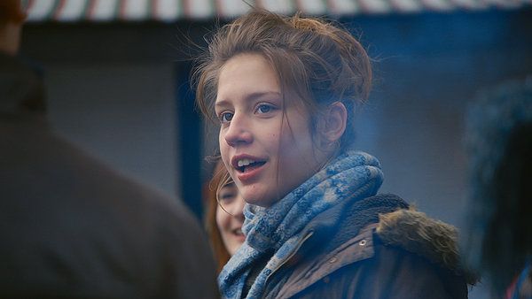 Young woman with light skin talking, wearing a blue scarf and a jacket, outdoors.