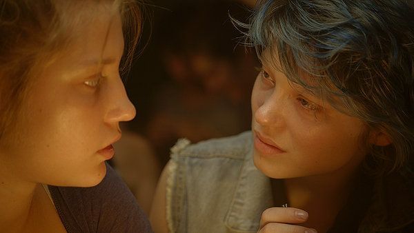Two young women facing each other. One has blue hair and looks sad. Dim lighting, close-up shot.