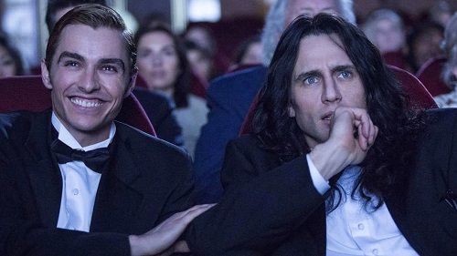 James Franco smiling next to Tommy Wiseau, both in tuxedos, watching a film in a theater.