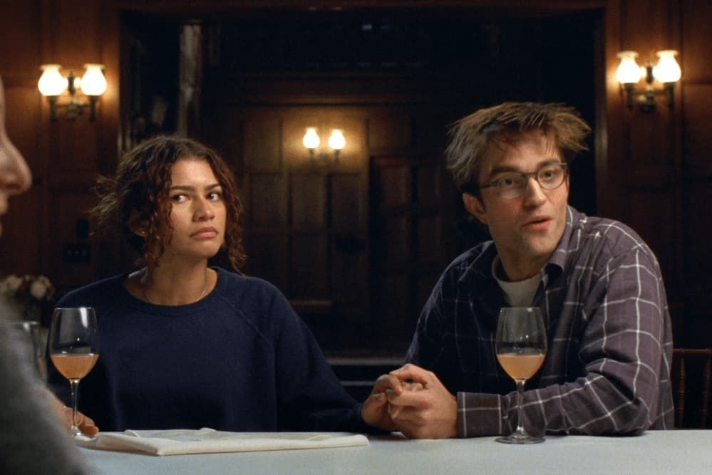 Two people seated at a table in a dim, wood-paneled room, each with a glass of white wine