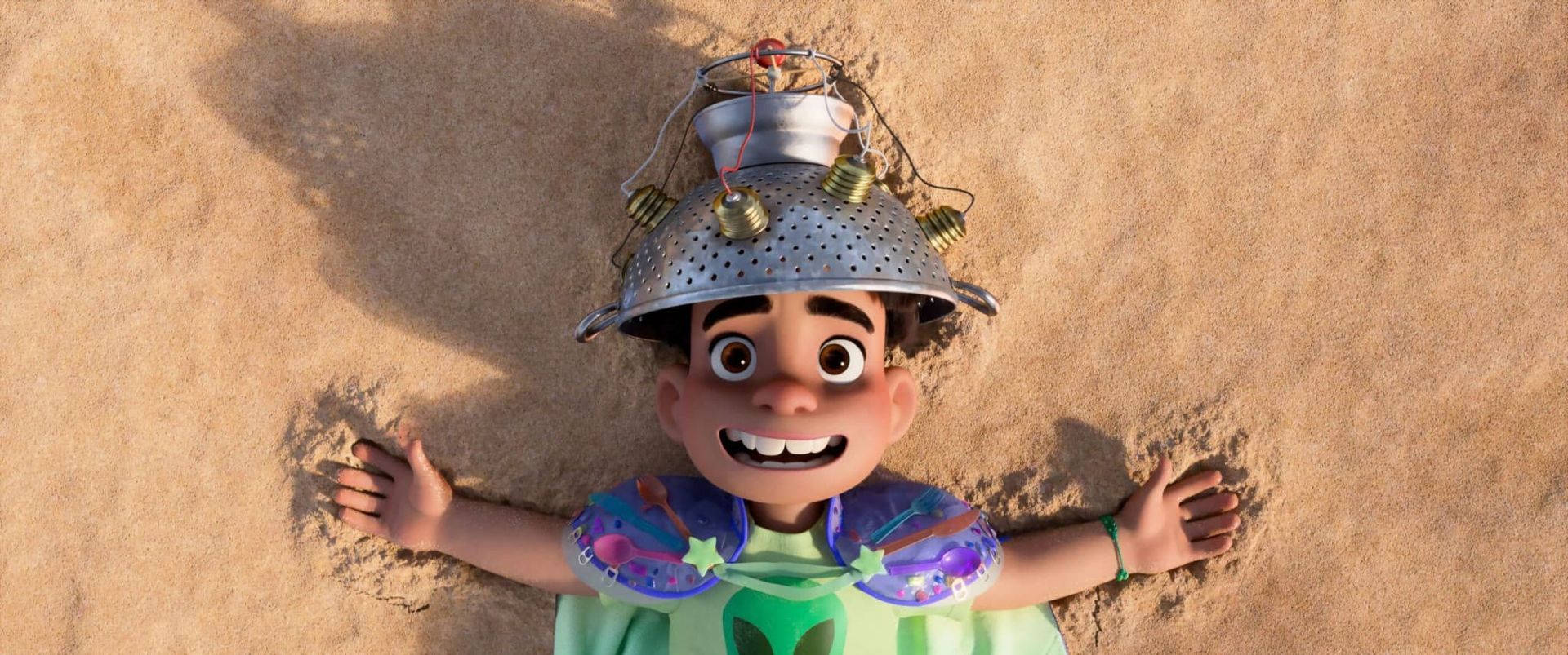 Smiling child lying in sand, wearing a colorful outfit and metal bucket hat with a toy on top