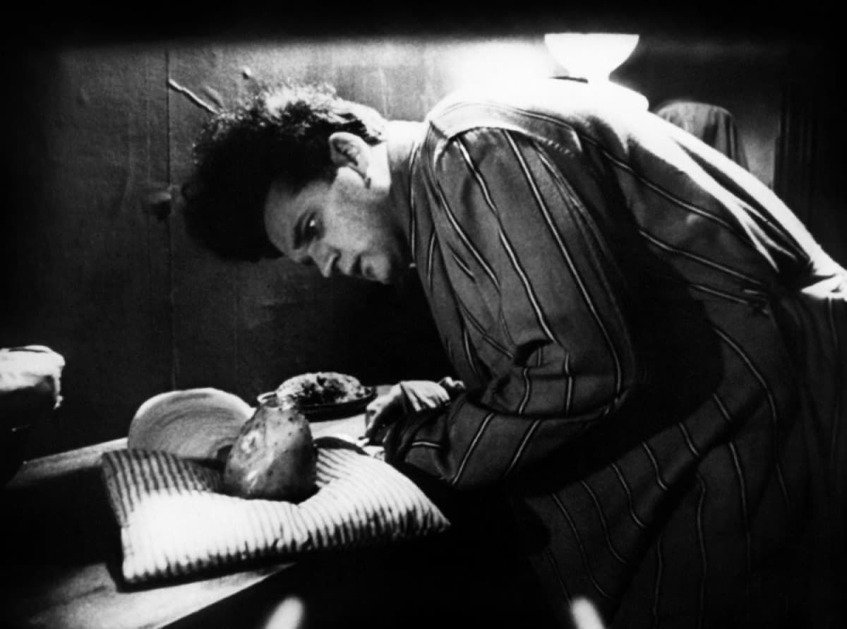 Black-and-white scene of a man leaning over a person lying in bed, looking down with concern.