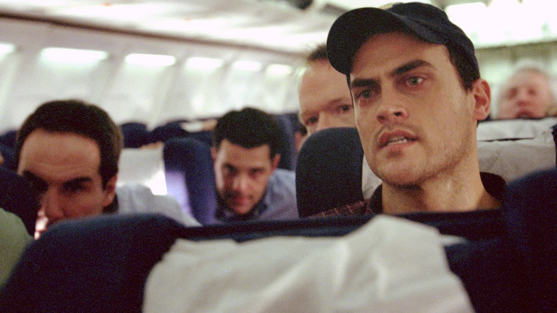 Passengers seated on a bus, with a man in a black cap in the foreground looking ahead