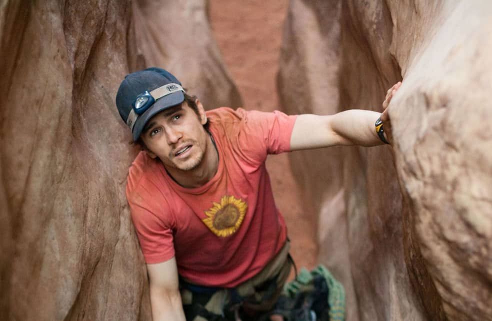 Man trapped in narrow canyon, dirt covered, struggling to climb, wearing red shirt and cap.