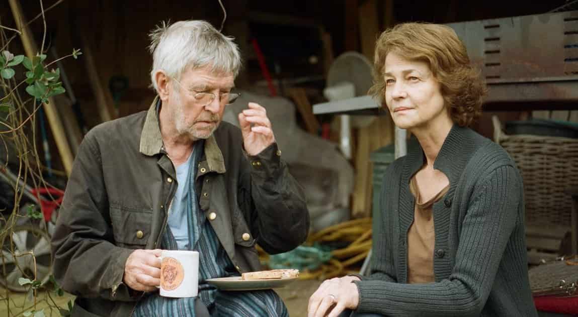 An older man and woman sit outside; man eats, woman looks on. They are in front of a cluttered shed.