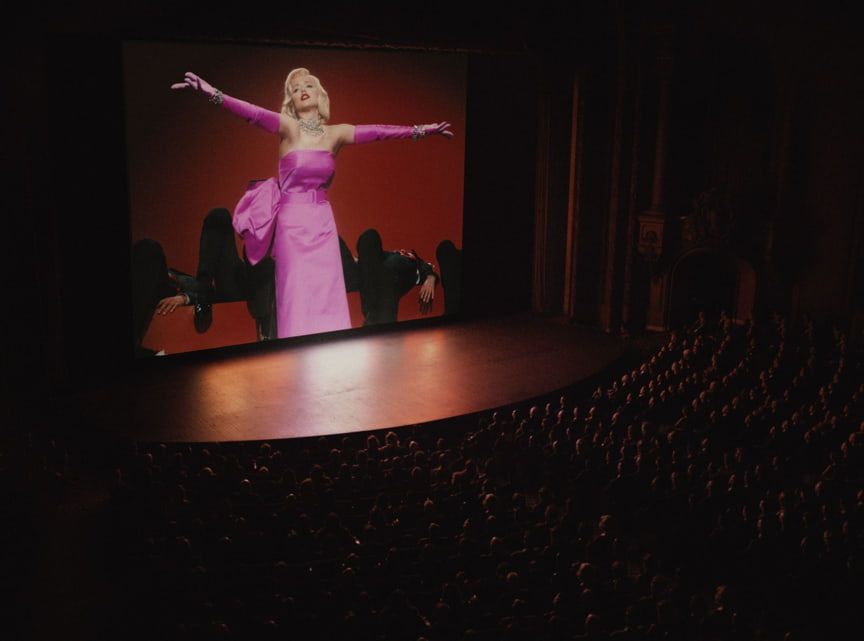 Blonde woman in pink dress on a large screen; stage set; audience in the dark.