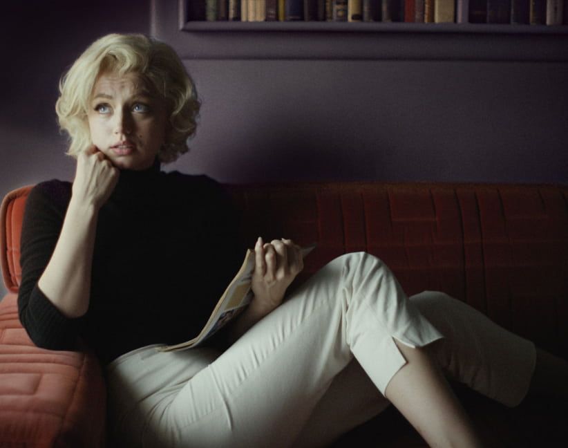 Ana de Armas as Marilyn Monroe, seated, gazing pensively, holding paper, in a room with books.