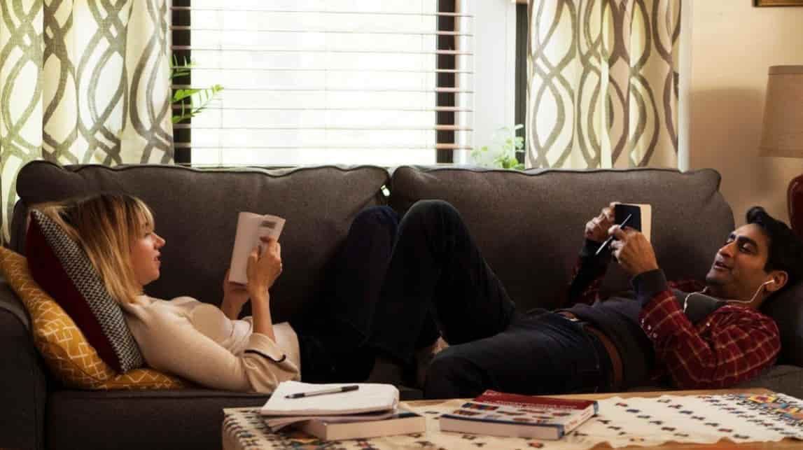 Woman reading a book, man playing video game on a couch. Sunlight streams through a window.