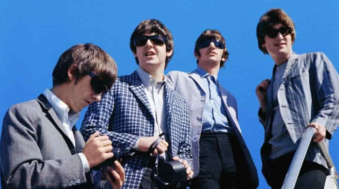 The Beatles in sunglasses and jackets against a blue sky, posing on a railing.