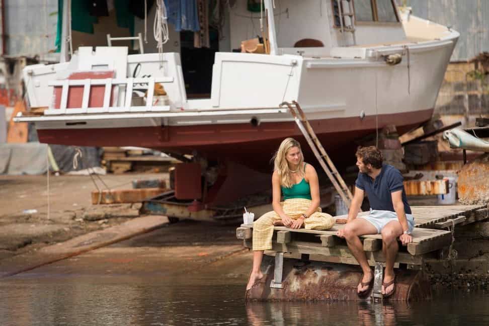 Couple sitting on a dock, talking near a beached boat. Bright clothes, sunny day, boatyard setting.