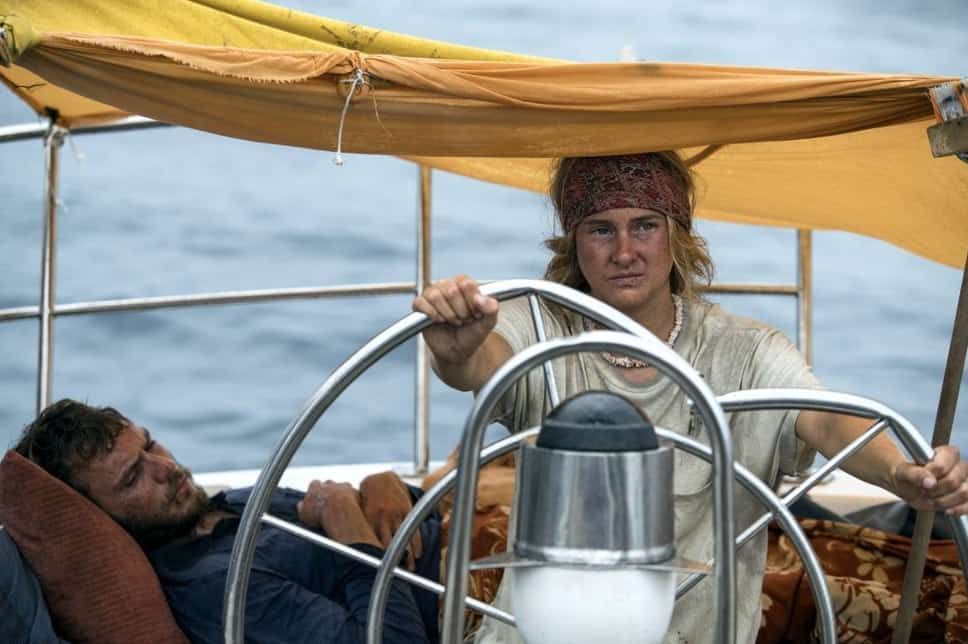 Woman steers sailboat, man rests. Overcast skies.
