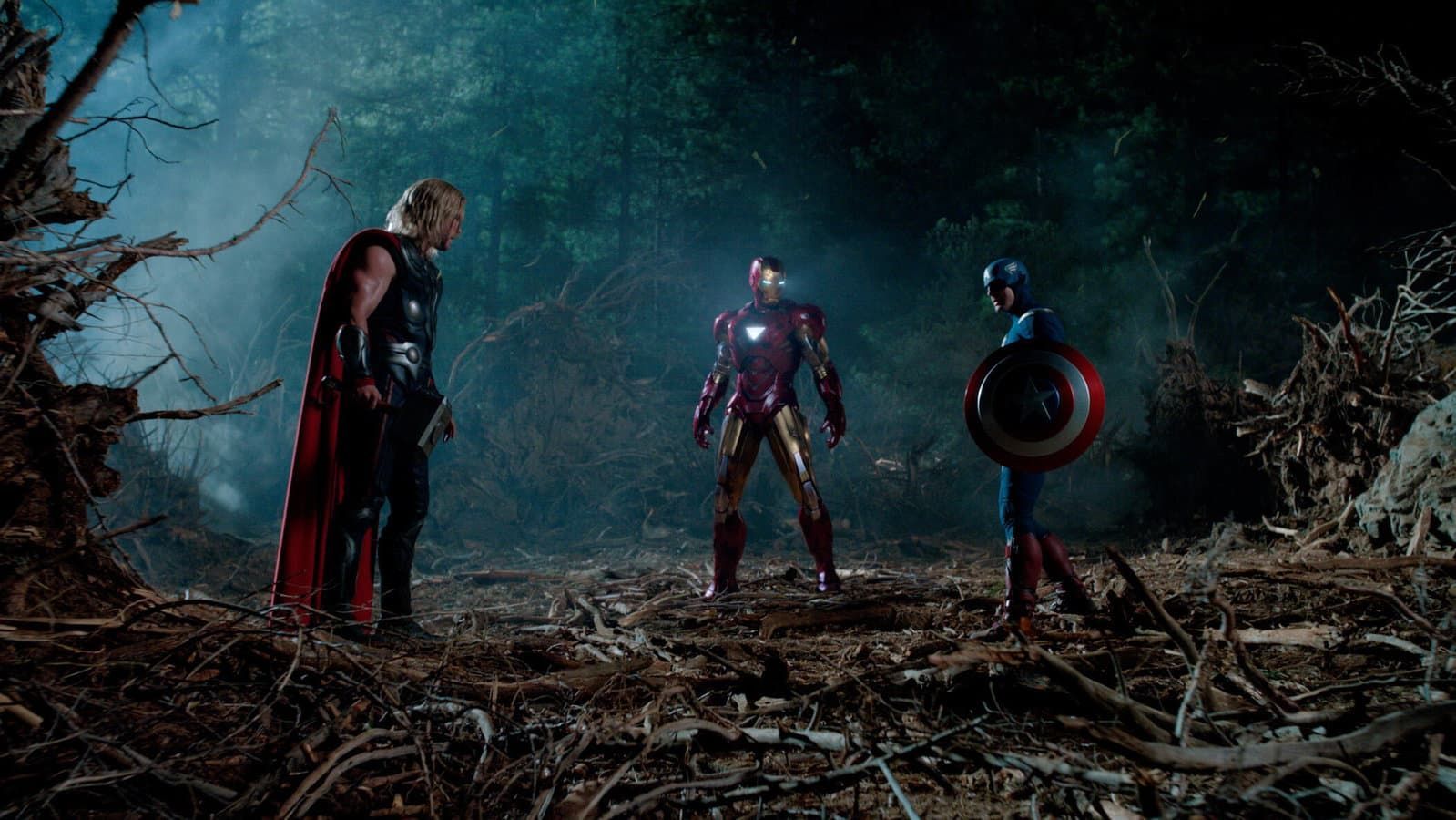 Three superheroes stand in a dark forest clearing, facing each other under blue mist.