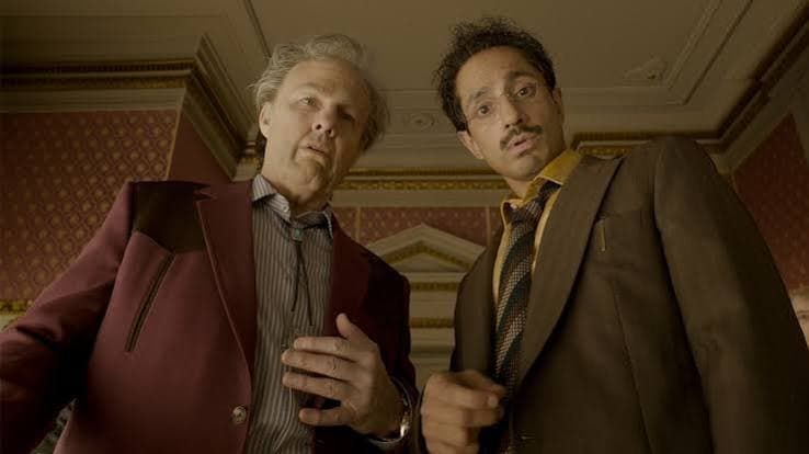Two men in suits lean over, looking down in a patterned room with pink wallpaper.
