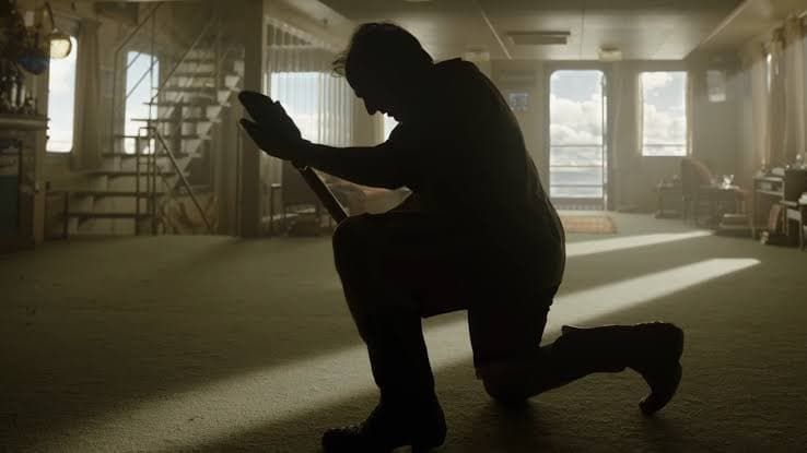 Silhouetted person kneeling in a sunlit empty room, holding a small object with one arm raised.