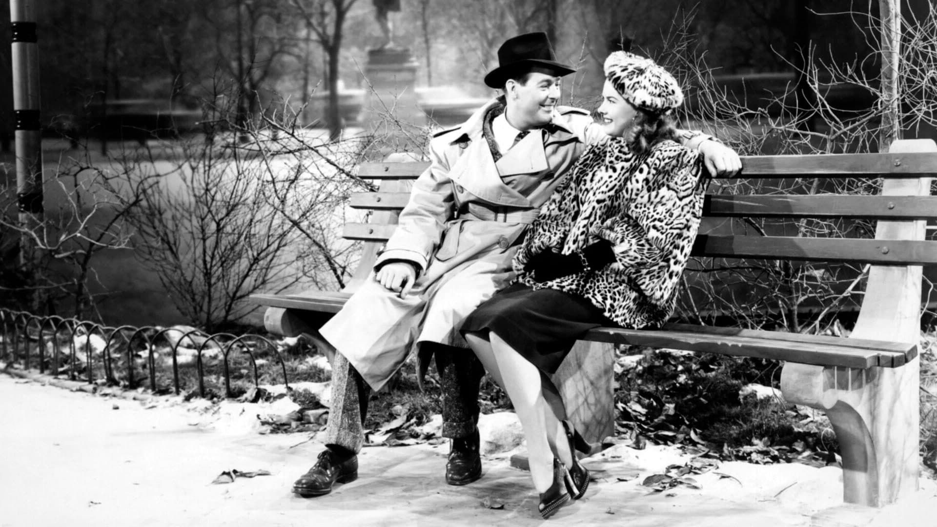 Two people in winter coats sit on a park bench, smiling and leaning toward each other in the snow.