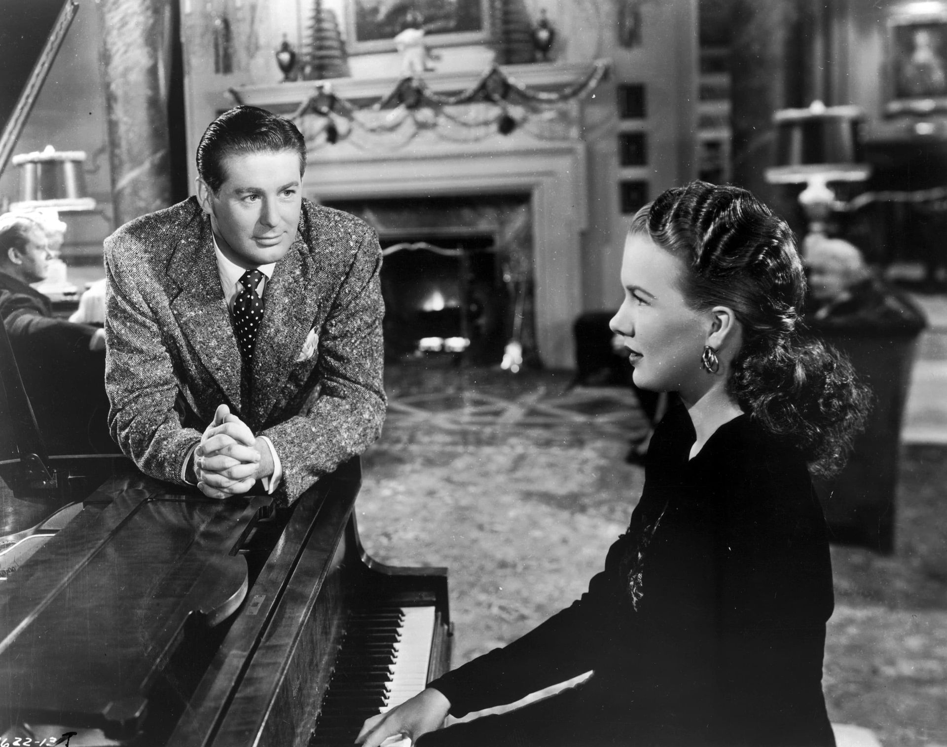 Black-and-white scene of a man leaning on a piano, talking to a woman seated beside it in a living room.