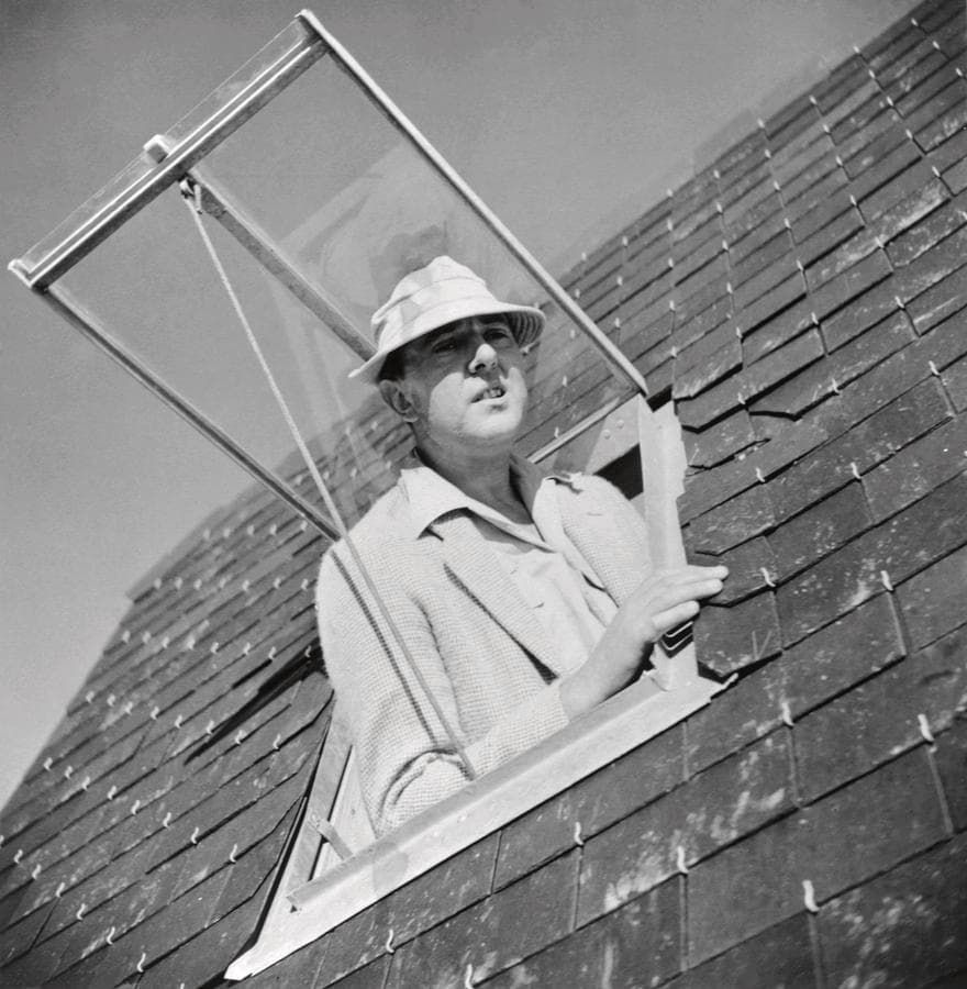 Man peeking from a roof skylight, wearing a hat and glasses, with a metal frame behind him.