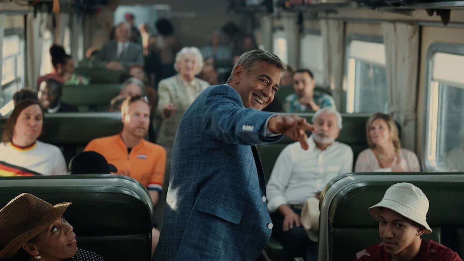 Man smiling and pointing while speaking on a bus, with seated passengers around him