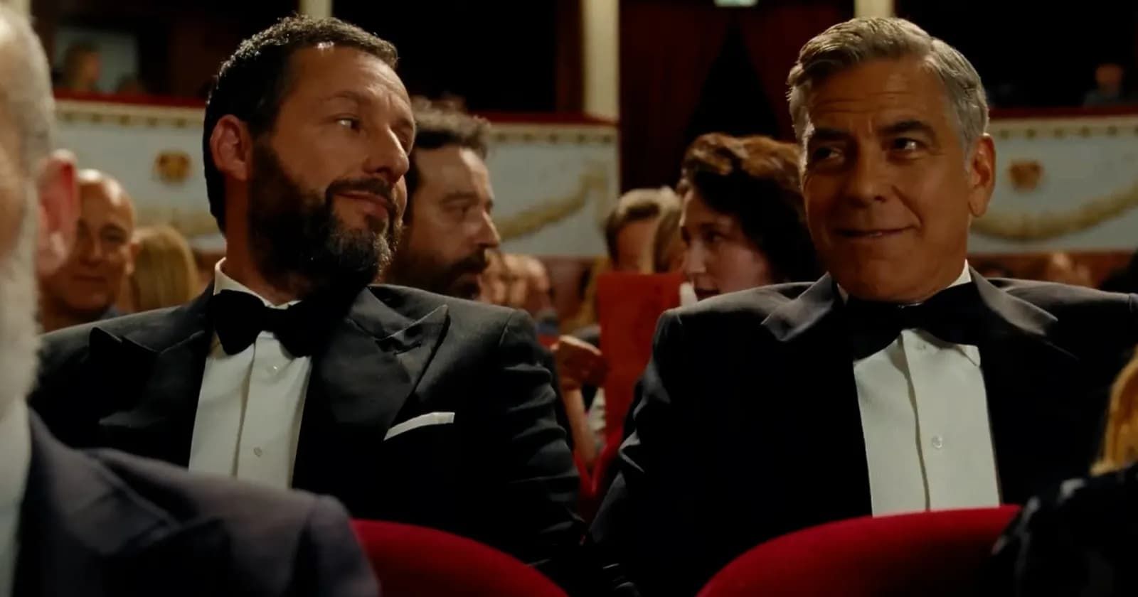 Two men in tuxedos seated in a theater, one smiling at the other.