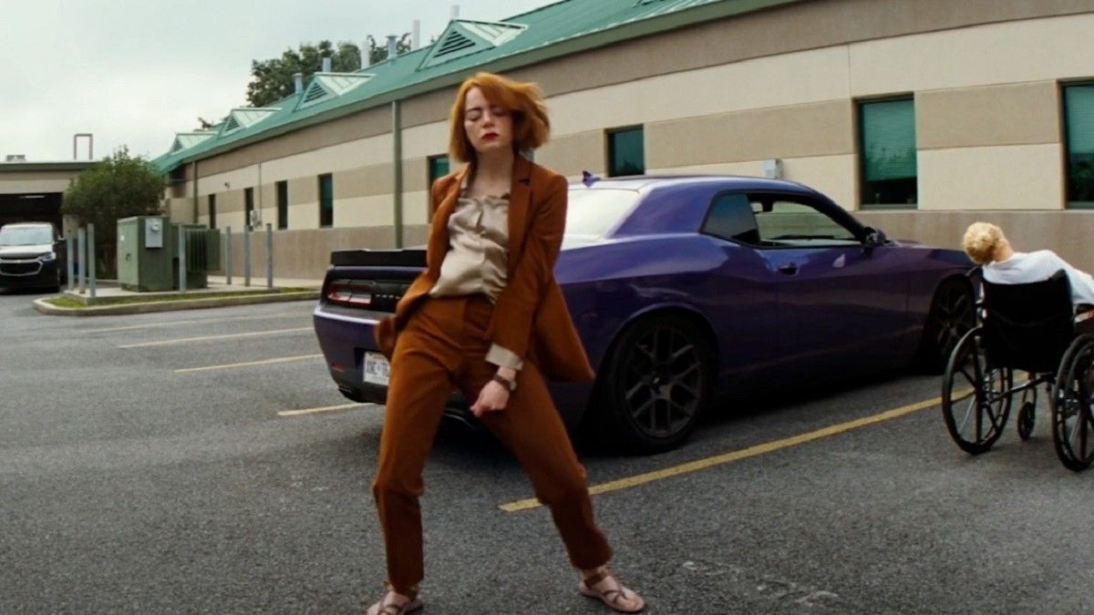 Woman in orange suit dances in parking lot near purple car and person in wheelchair.