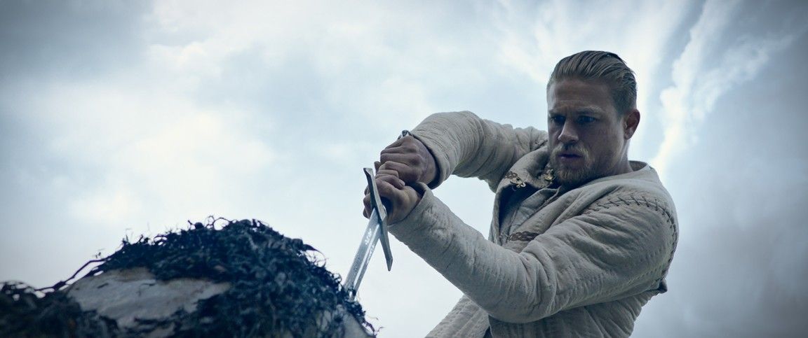 Man in white sweater thrusts sword into object, against cloudy sky.