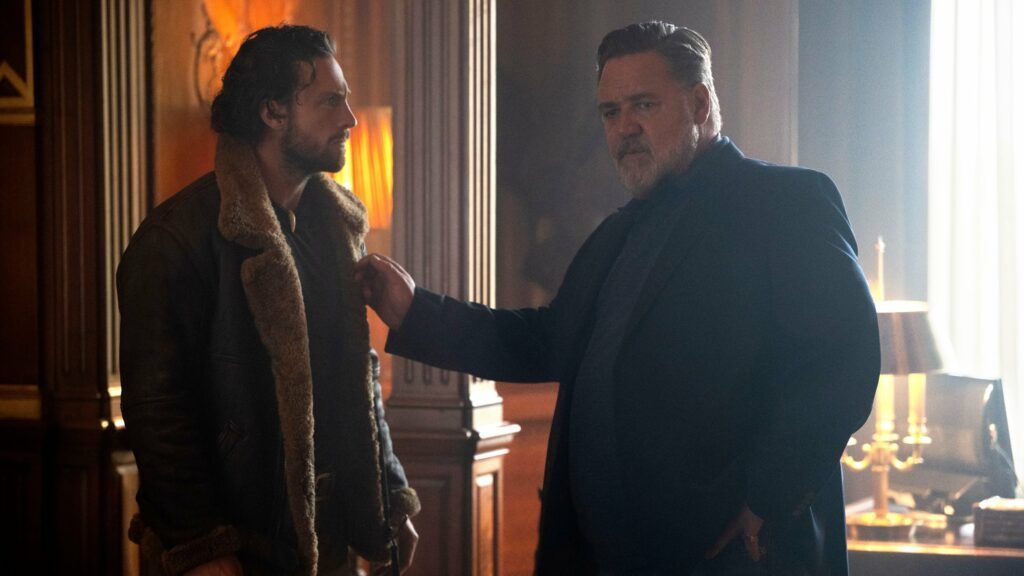 Two men in a room; older man touches younger man's jacket. Dimly lit with wood paneling.
