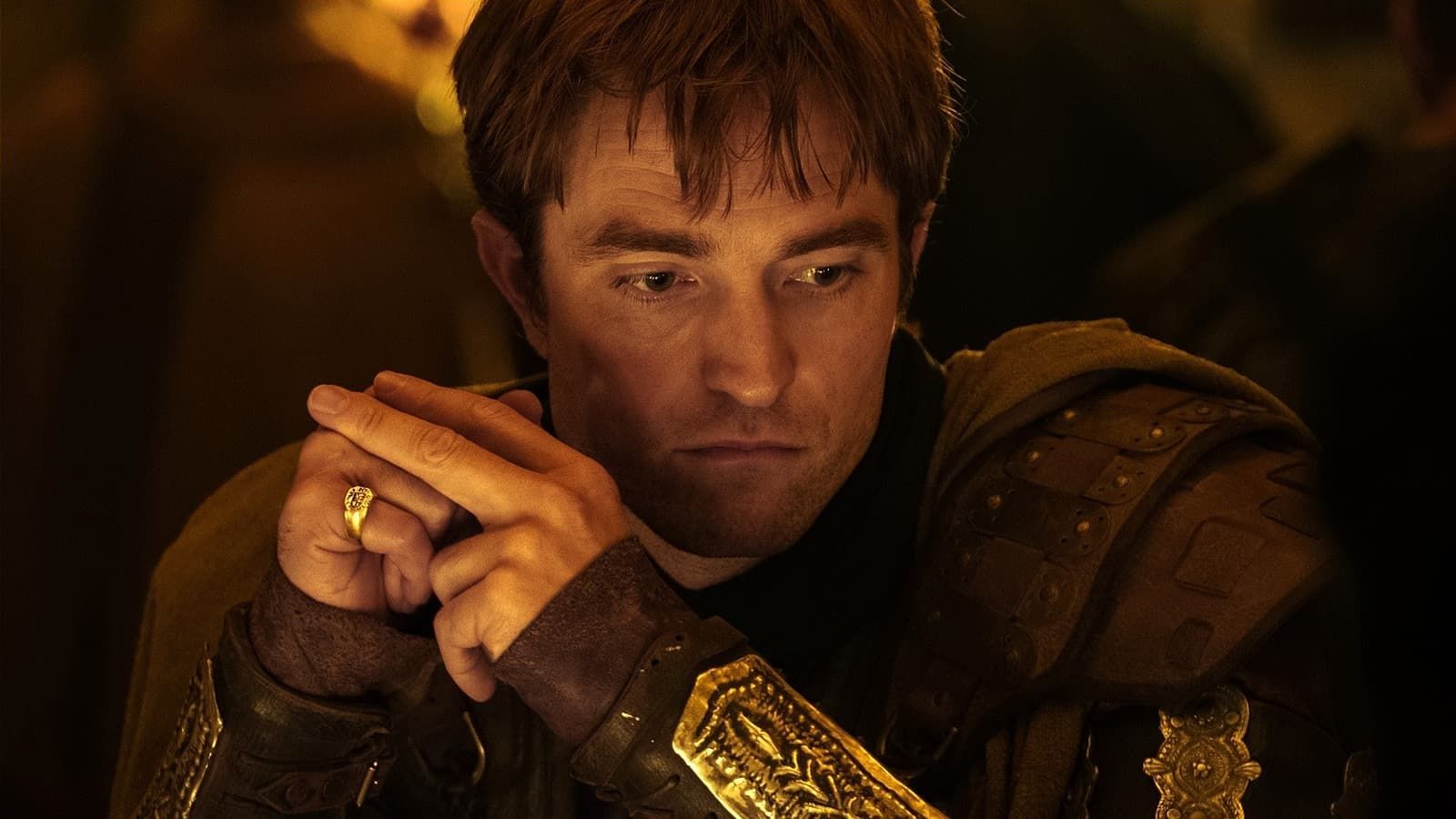 A young man in ornate armor sits in warm firelight, hands clasped and looking down thoughtfully.