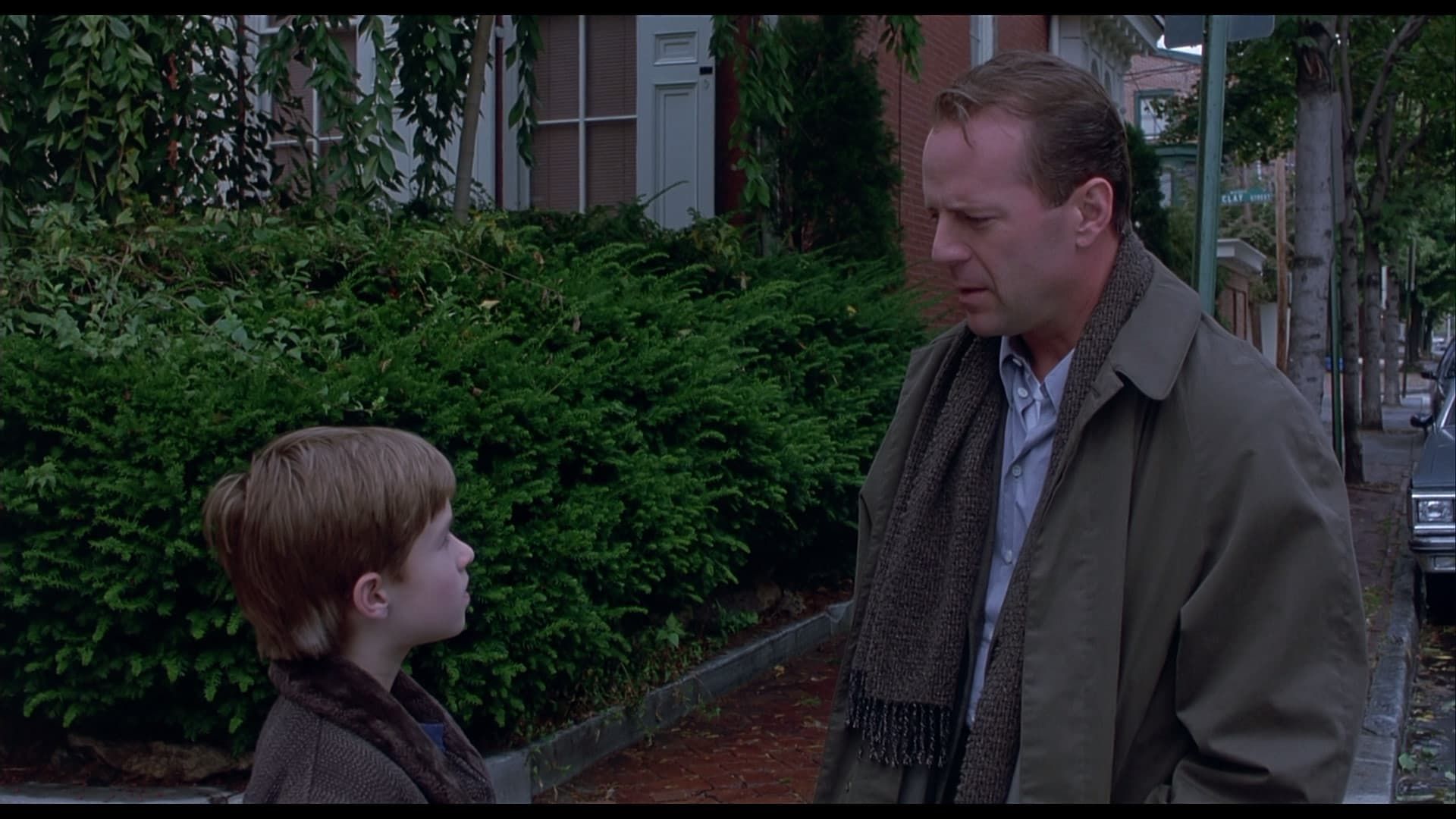 Man and child talking on a sidewalk beside hedges on a rainy street.