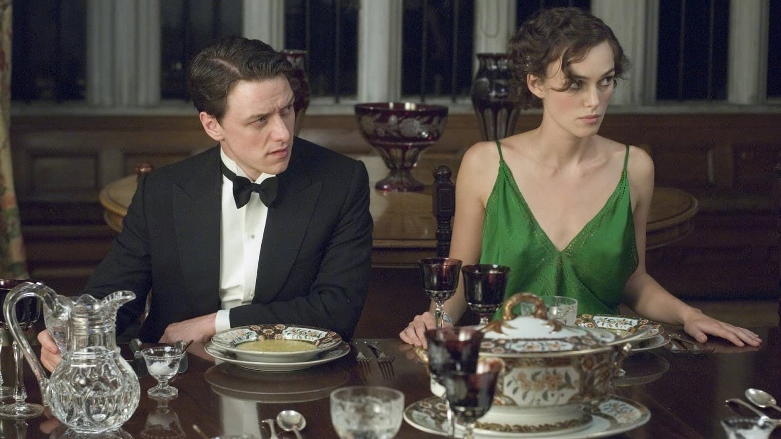 Two people in formal attire sit at a dining table, looking in opposite directions.
