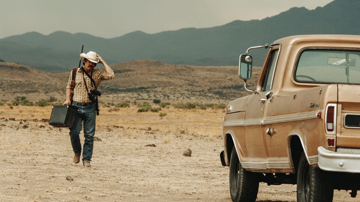 Person walking across a dry desert road beside a tan pickup truck, with mountains in the background