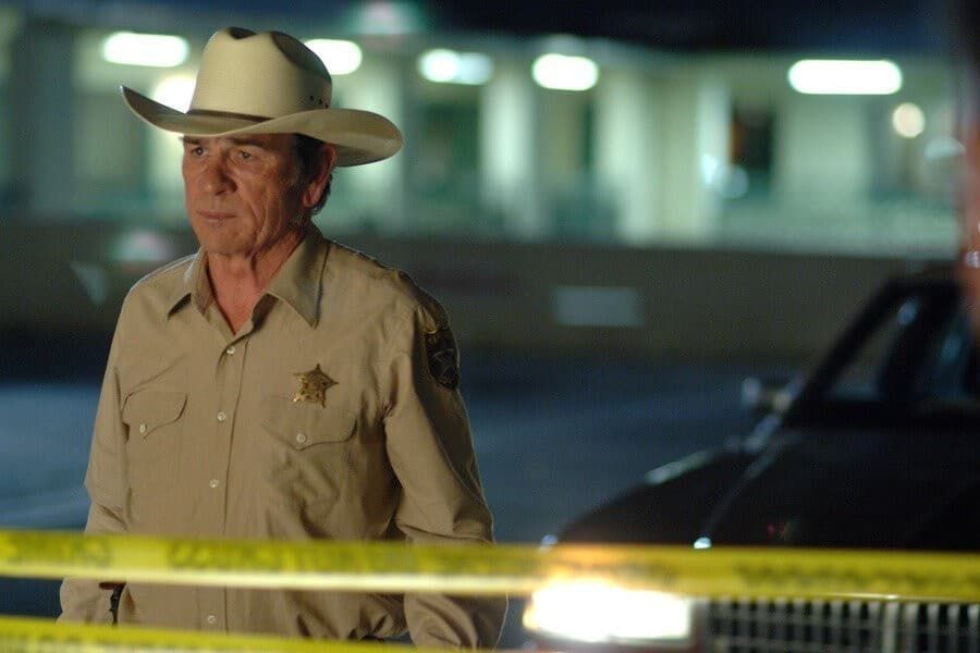 Sheriff in tan uniform and white hat stands behind yellow tape at a nighttime crime scene