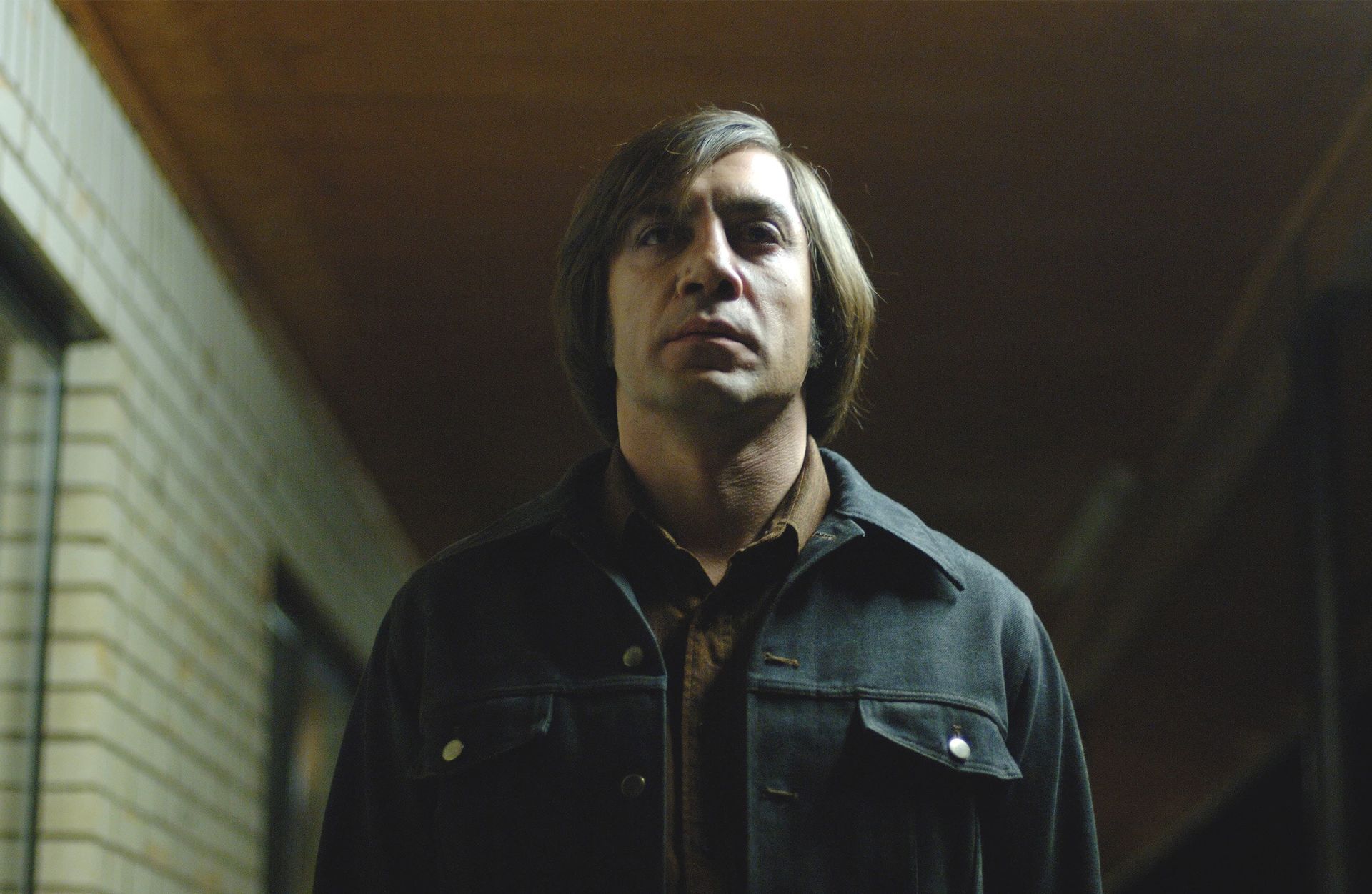 A serious man in a dark jacket stands in a dim hallway, lit from above.