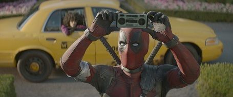 Deadpool holding a camera in front of a yellow car with a driver visible inside.
